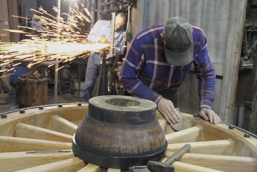 Calvin Roorda sands a spoke on a huge wheel while Dave Engel grinds a brake ratchet at the Engel Coach Shop in Joliet. Click on the arrow at top right for more photos. (Ed Kemmick/Last Best News)